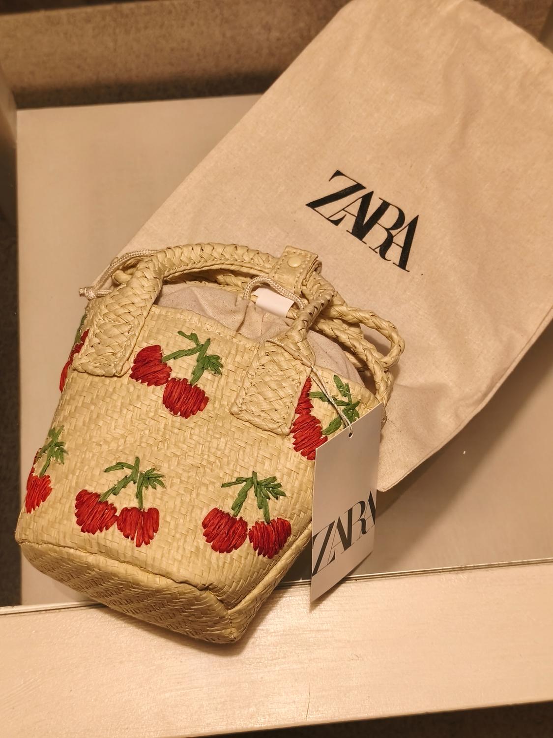 Straw bag with red cherry embroidery and Zara tag, on a fabric bag with the Zara logo.
