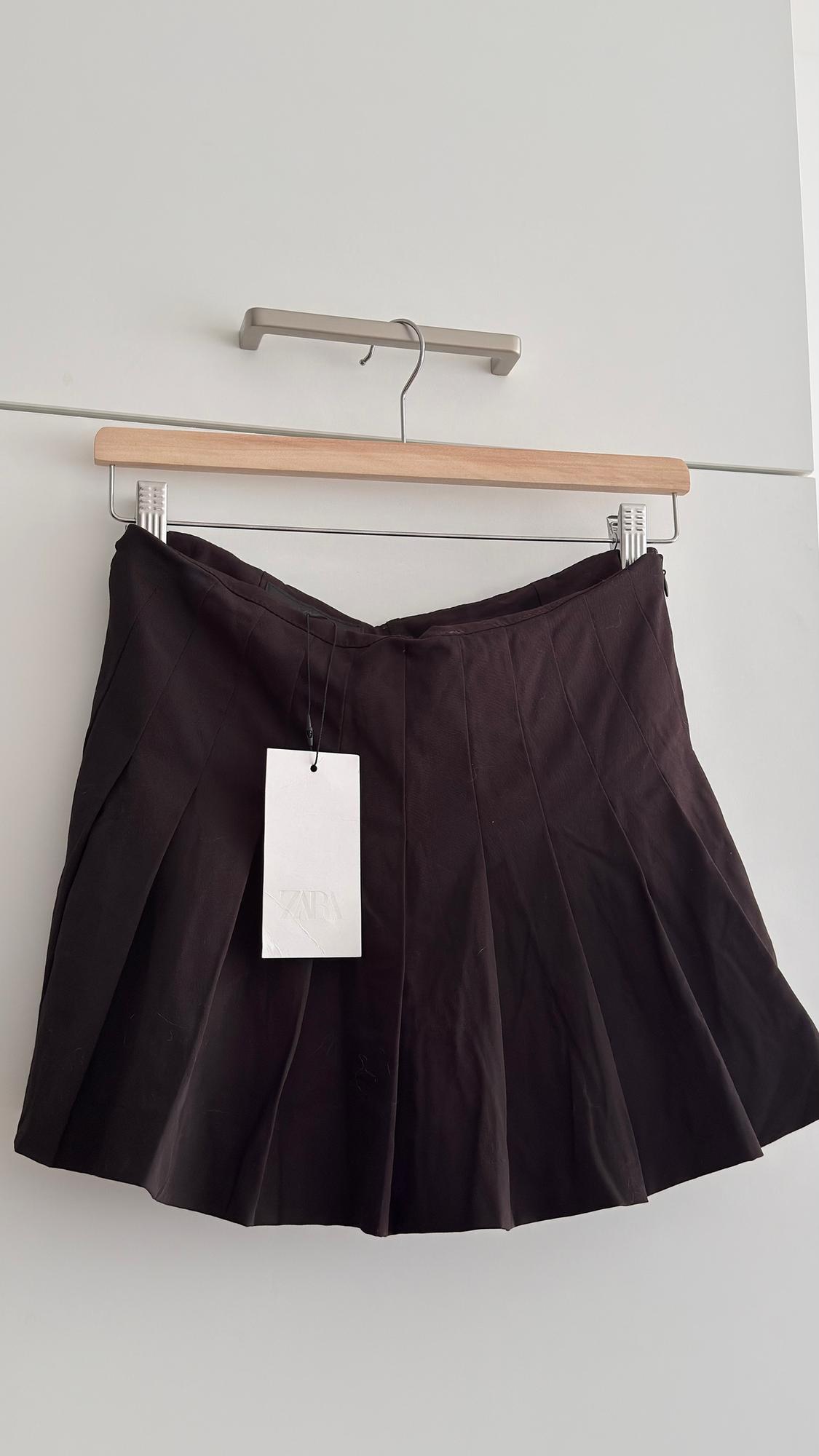 Black pleated skirt hanging on a wooden hanger with a white "ZARA" tag, against a white background.
