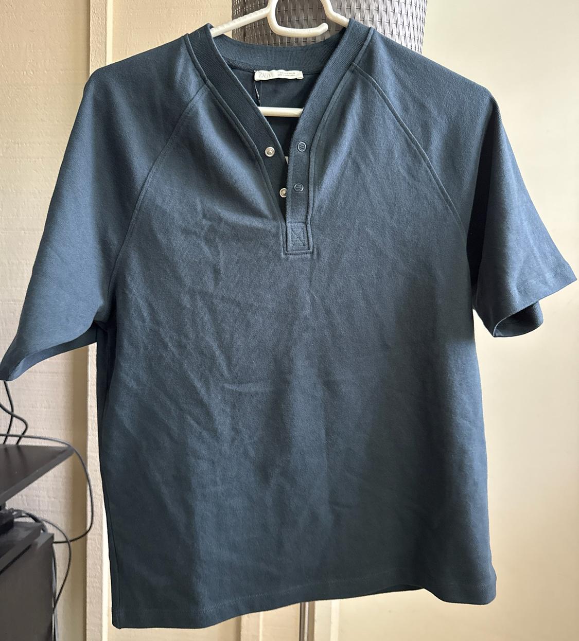 Dark blue short-sleeved T-shirt with a buttoned collar, hanging on a white hanger indoors.
