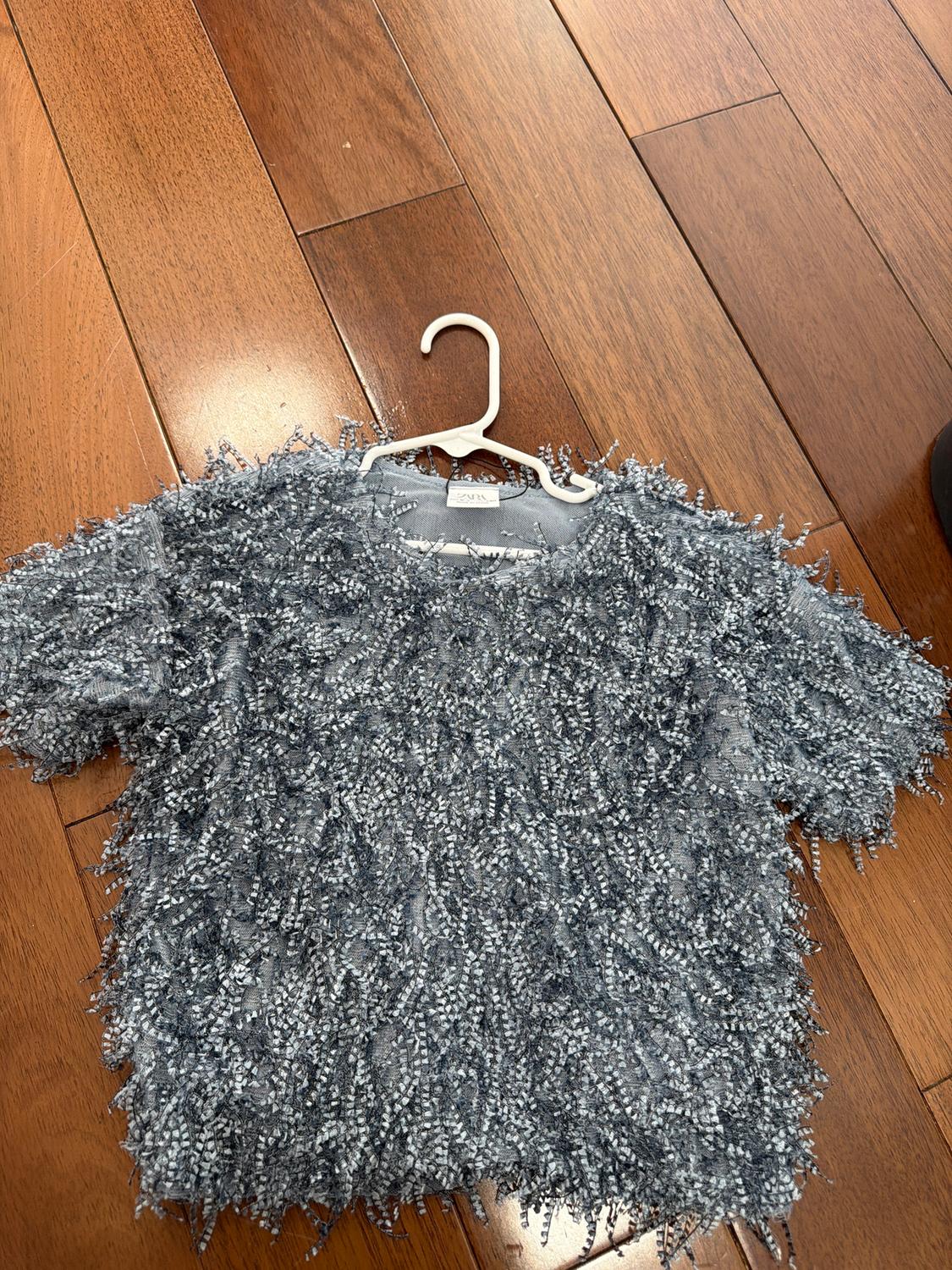 Textured gray t-shirt with fringe on a white hanger, placed on a brown wooden floor. Visible tag: "ZARA".