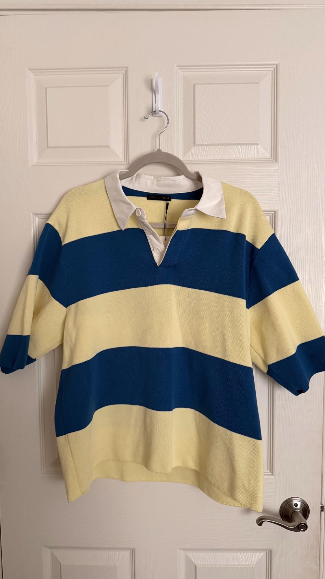 Short-sleeved polo shirt with broad yellow and blue stripes and a white collar, hanging on a hanger on a white door.