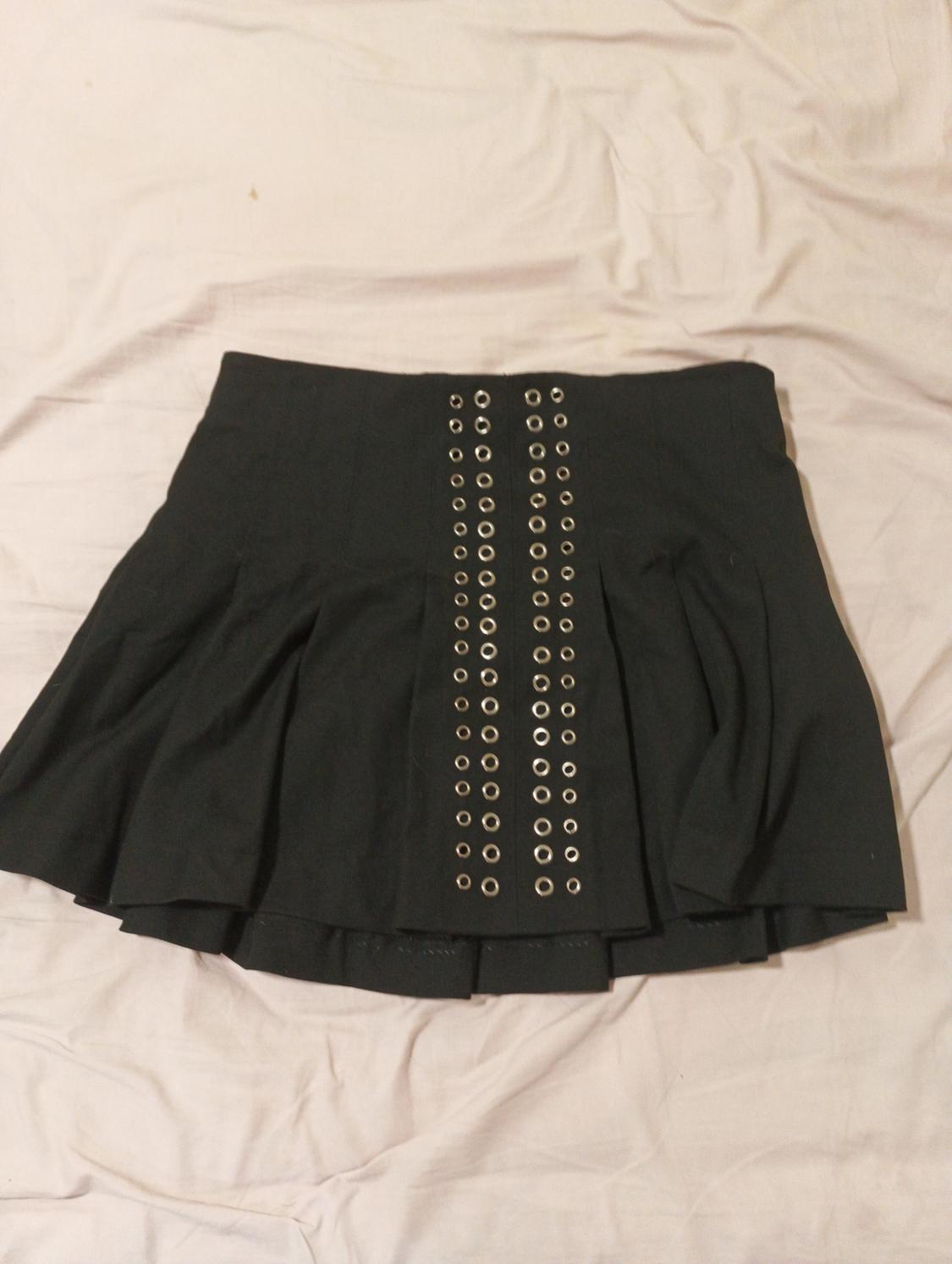 Black skirt with metallic eyelets in two vertical rows on a light-coloured fabric. Pleated and modern design.