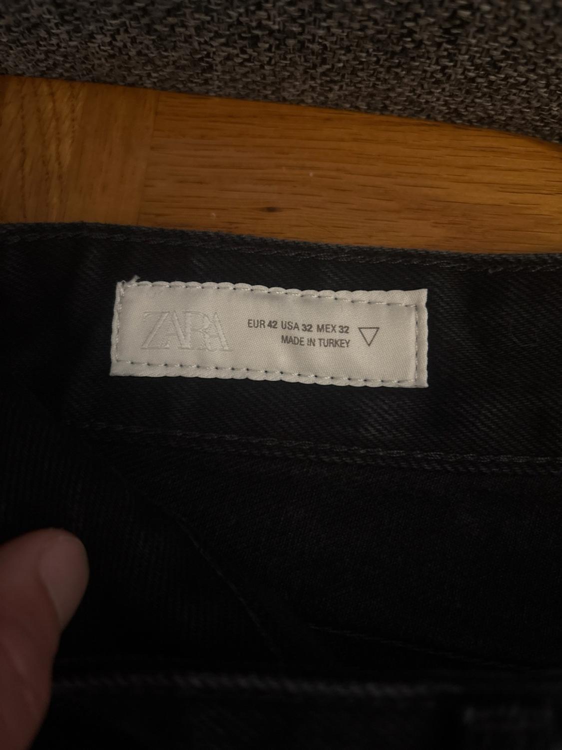 Close-up of a white label stitched onto black fabric. Text: ZARA EUR 42 USA 32 MEX 32 MADE IN TURKEY.
