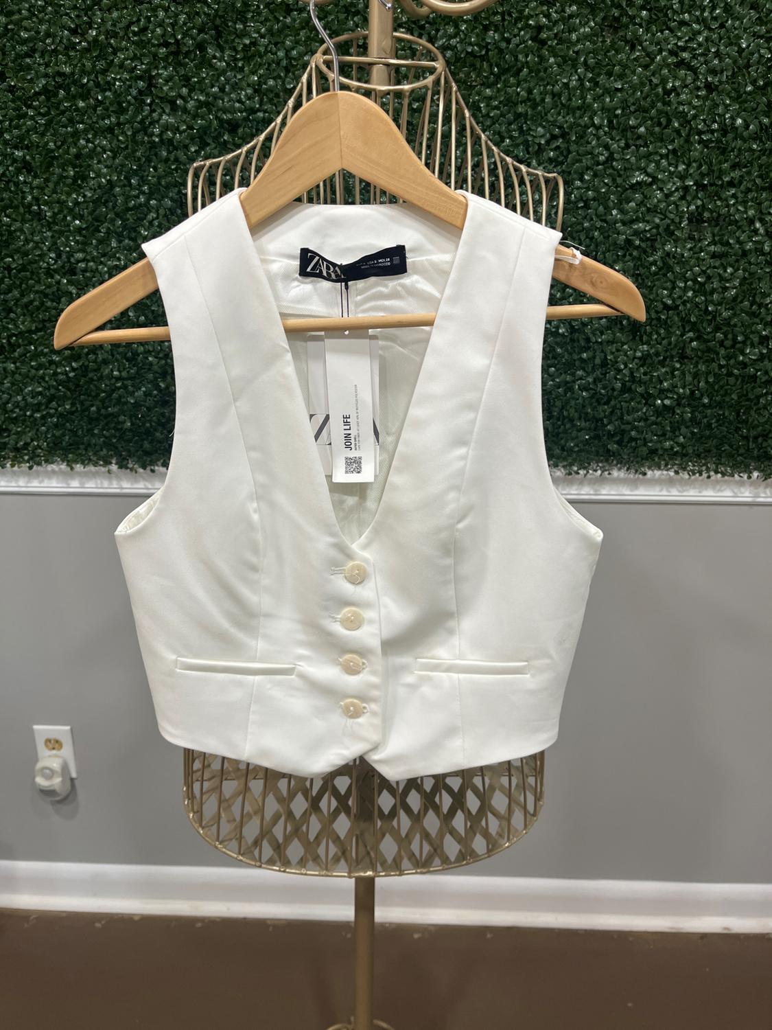 White button-up vest on a metal mannequin, hanging on a wooden hanger. Green wall background with leaf texture.