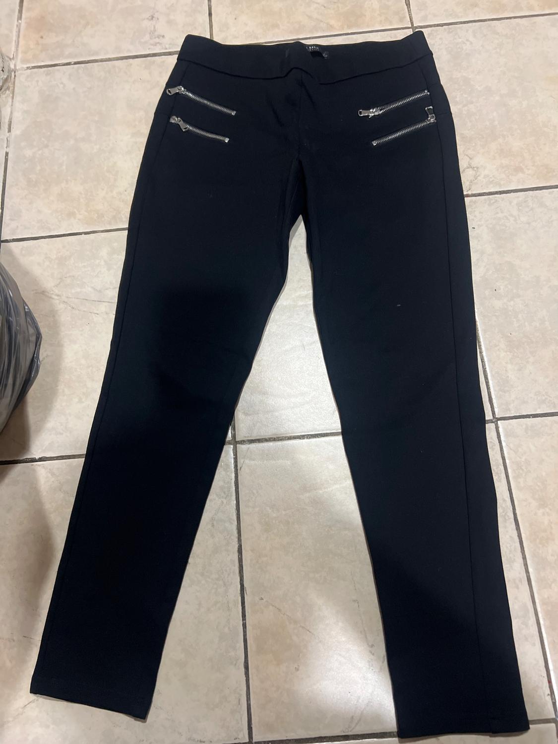 Black trousers with silver zips at the top, laid on a beige tiled floor.