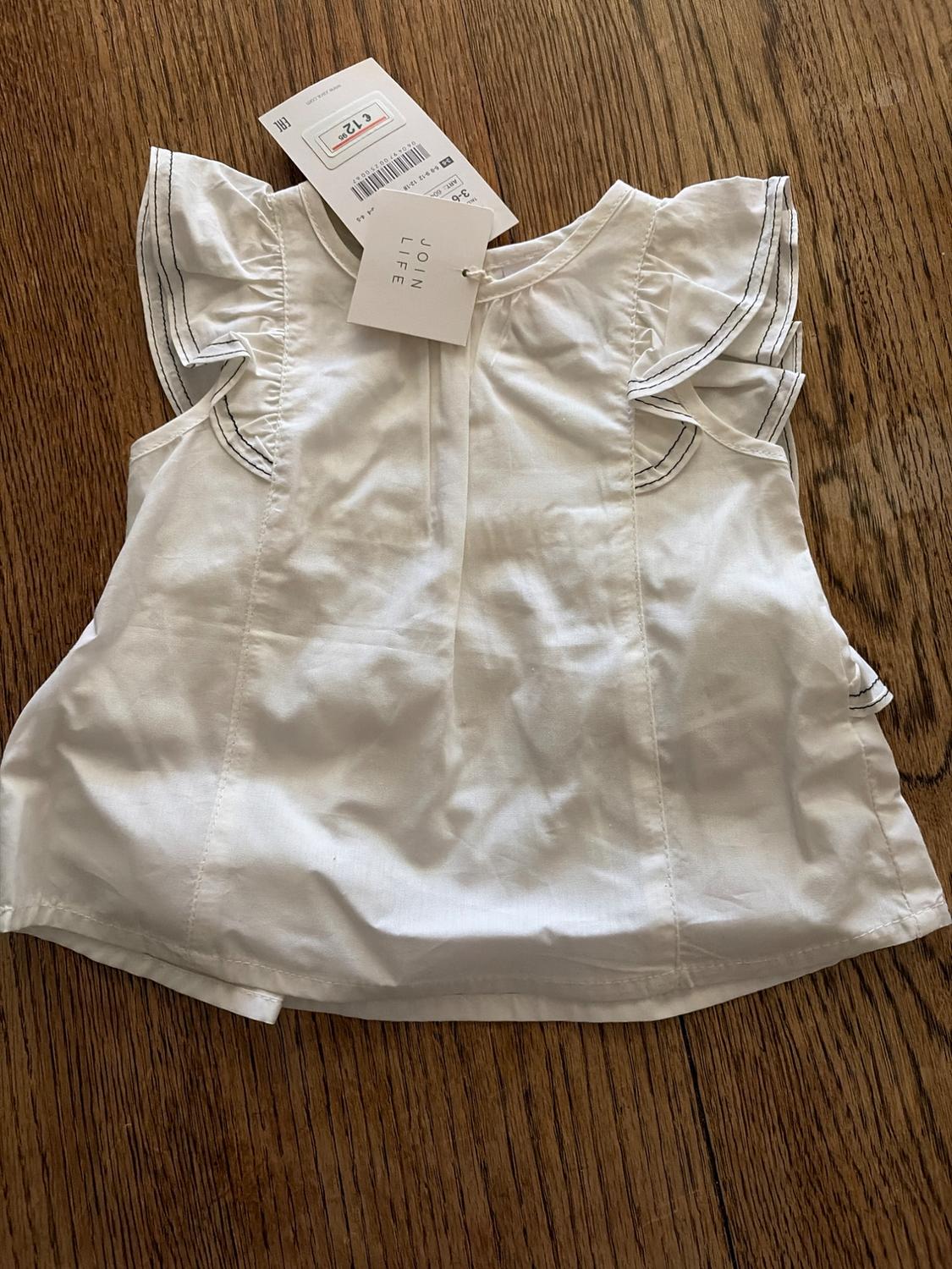 Girl's white blouse with frilled sleeves and black stitching, on a wooden surface. Visible label with text "JOIN LIFE".