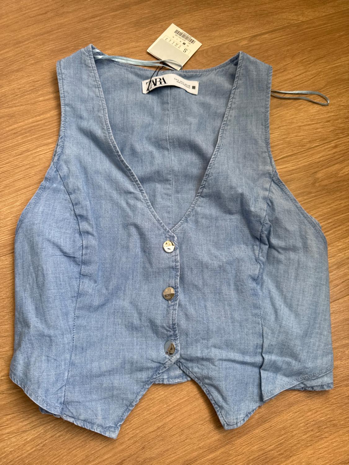 Light blue denim waistcoat with front buttons on wooden surface. Visible label with size S and ZARA brand.