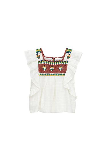 White blouse with ruffled sleeves and a multicolored crochet panel on the chest with a square neckline, on a white background.