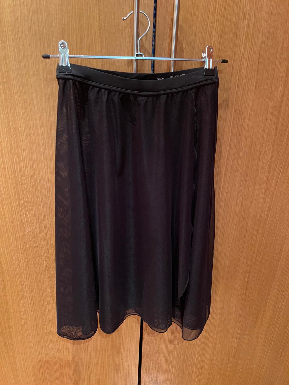 Flowing, semi-transparent black skirt hanging on a metal hanger in front of a wooden wardrobe. ZARA text on the waist.