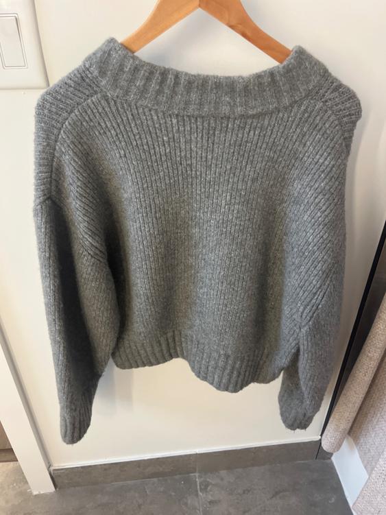 Gray chunky knit sweater hanging on a wooden hanger against a white wall, showing texture and simple design.