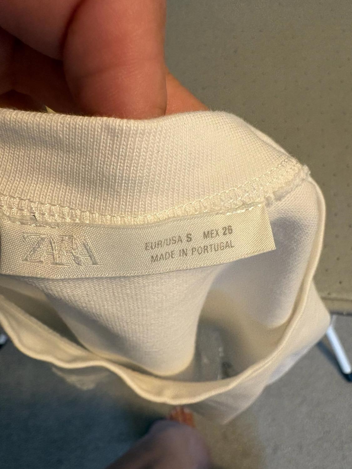 Close-up of a hand holding a white garment, showing the label with the text ZARA EUR/USA S MEX 26 MADE IN PORTUGAL.