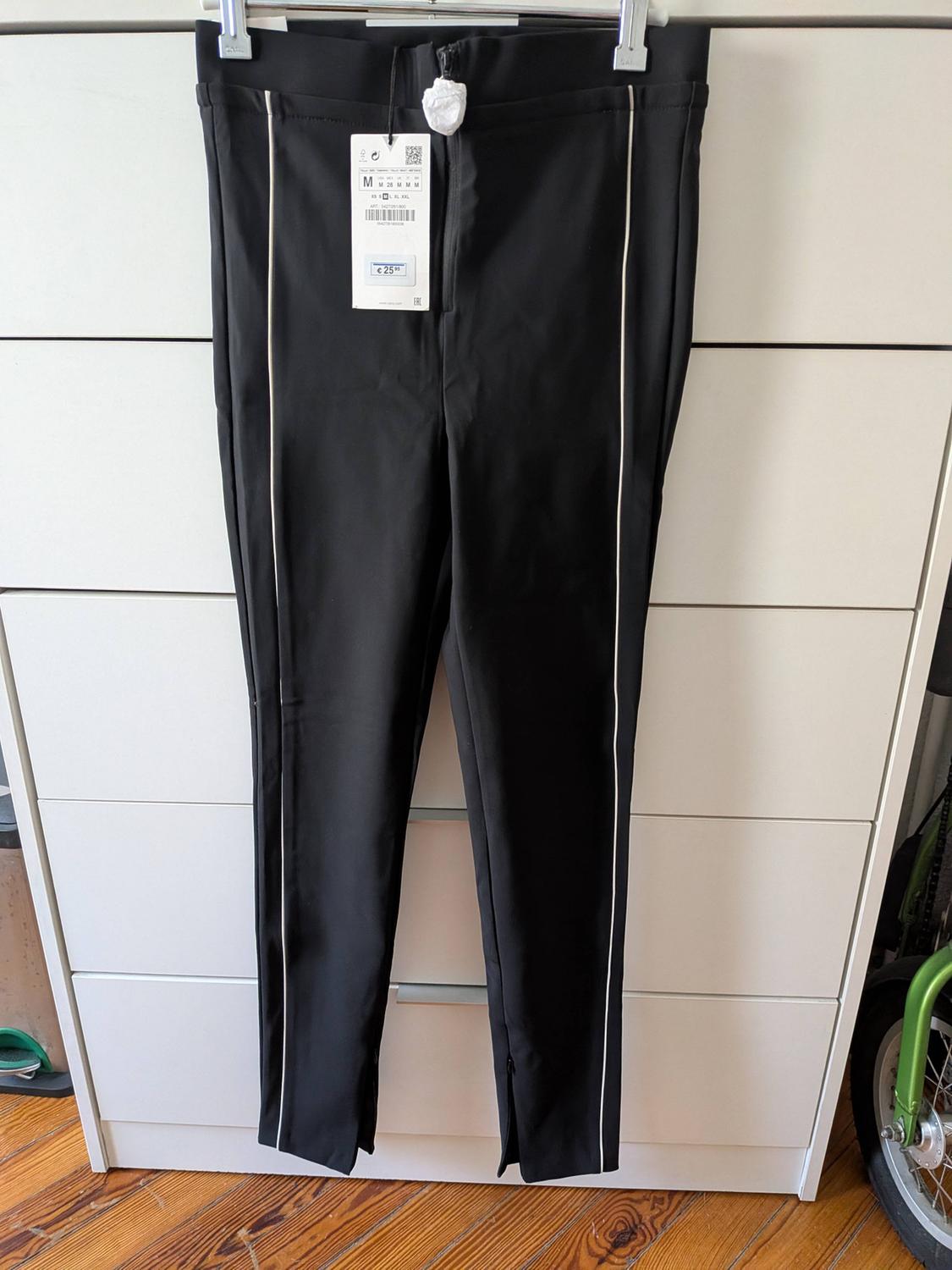 Black trousers with white side stripes hanging on a hanger in front of a white chest of drawers and a wooden floor. Label with text M € 25.