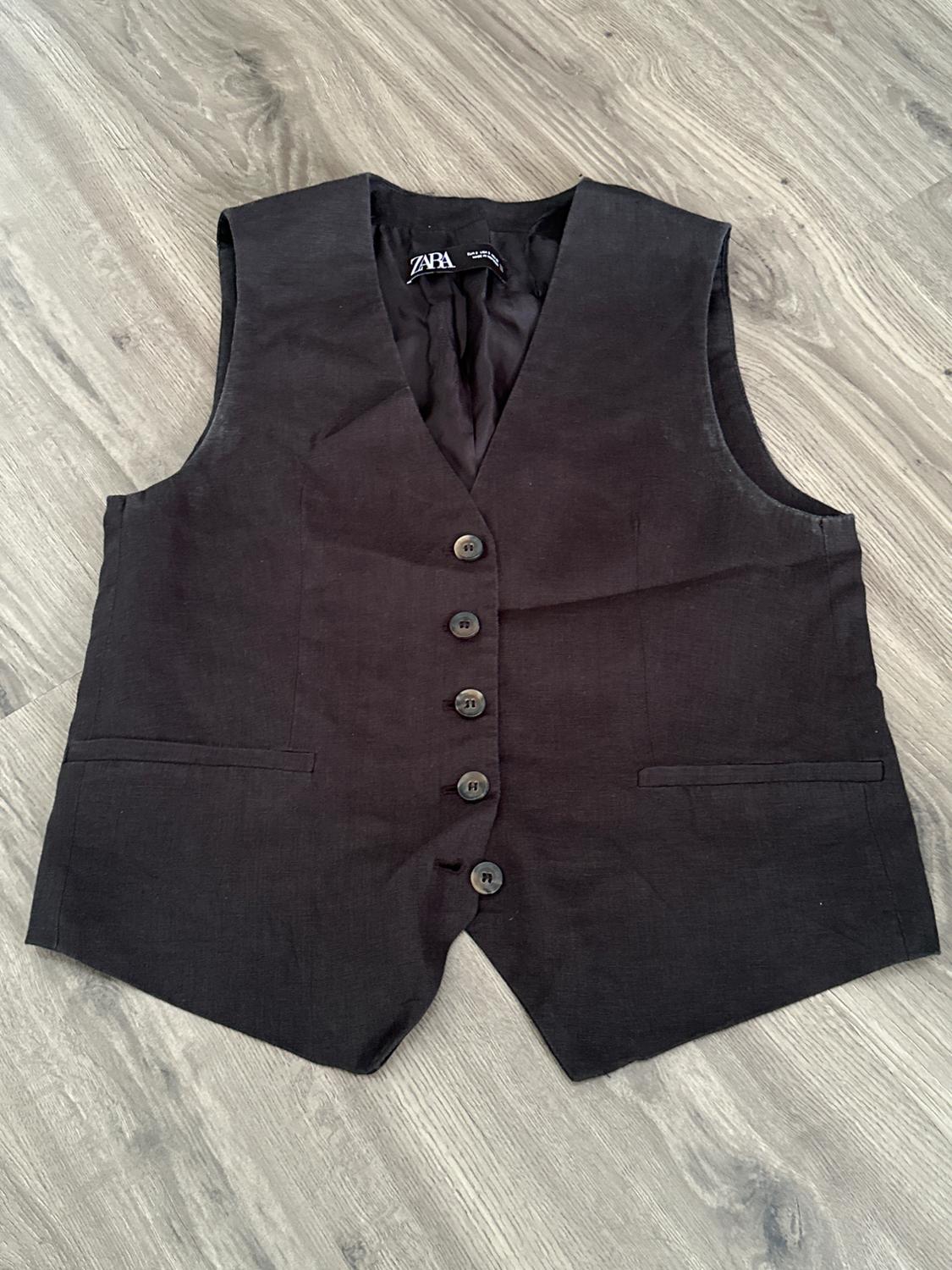 Black button-up waistcoat on a light wooden floor. Visible label with the brand "ZARA".