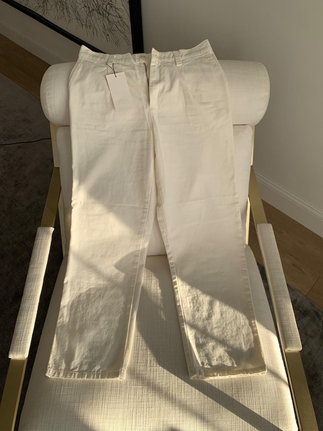 White trousers with a ZARA label on a white armchair with gold details. Natural light and shadows in a relaxed setting.