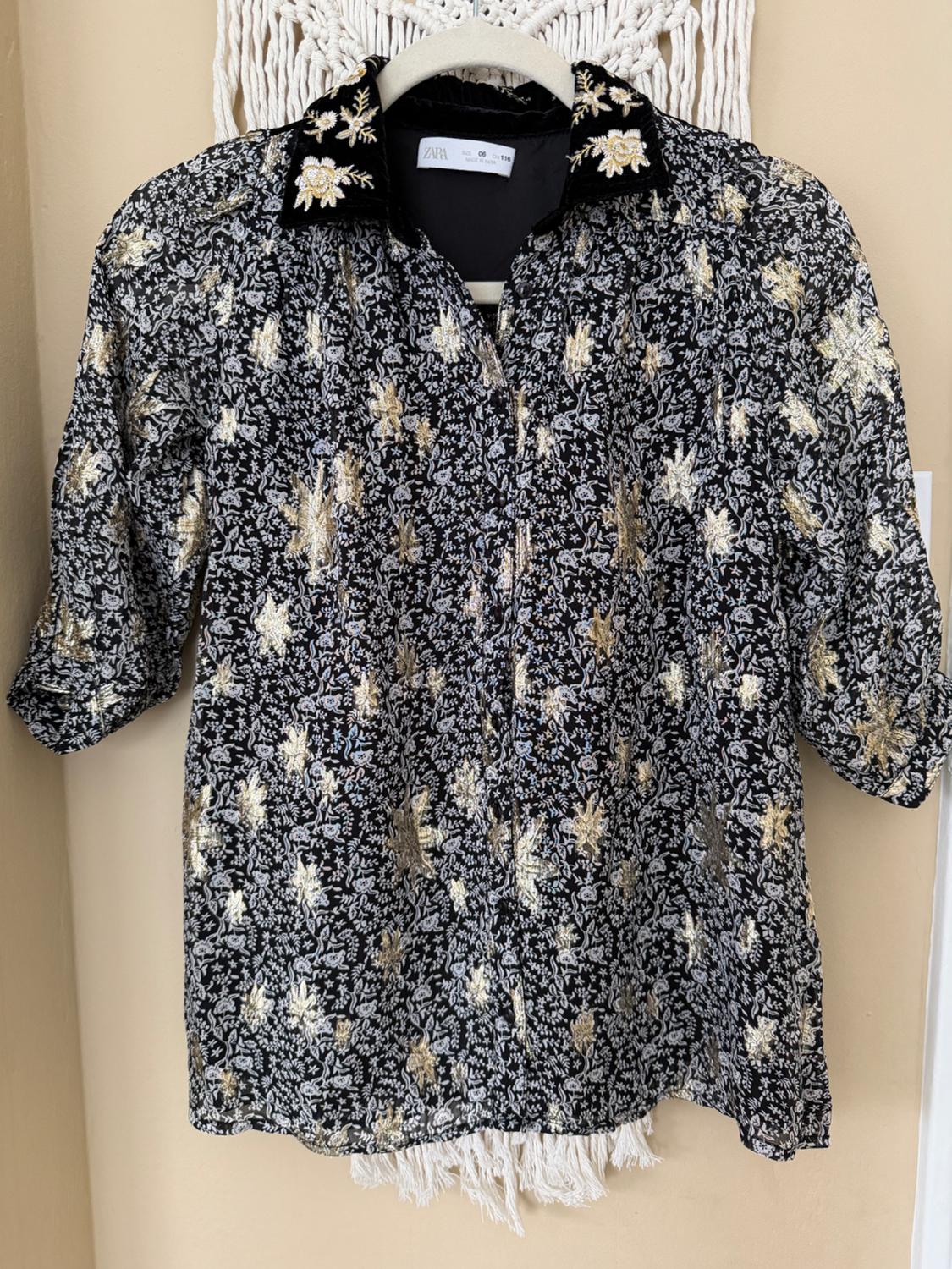 Blouse hanging with gold and black floral print, neck details. Visible tag with text "ZARA". Beige background with decorative fabric.