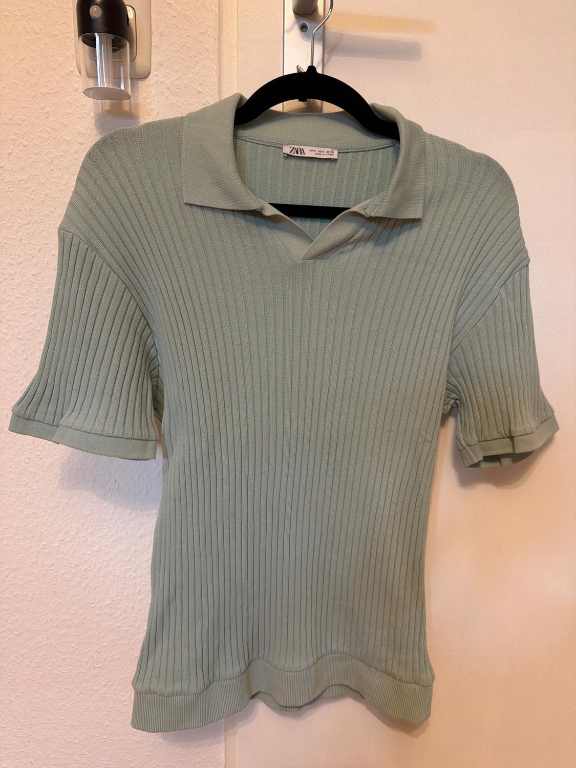 Light green ribbed knit polo neck jumper with short sleeves, hanging on a black hanger over a white door.