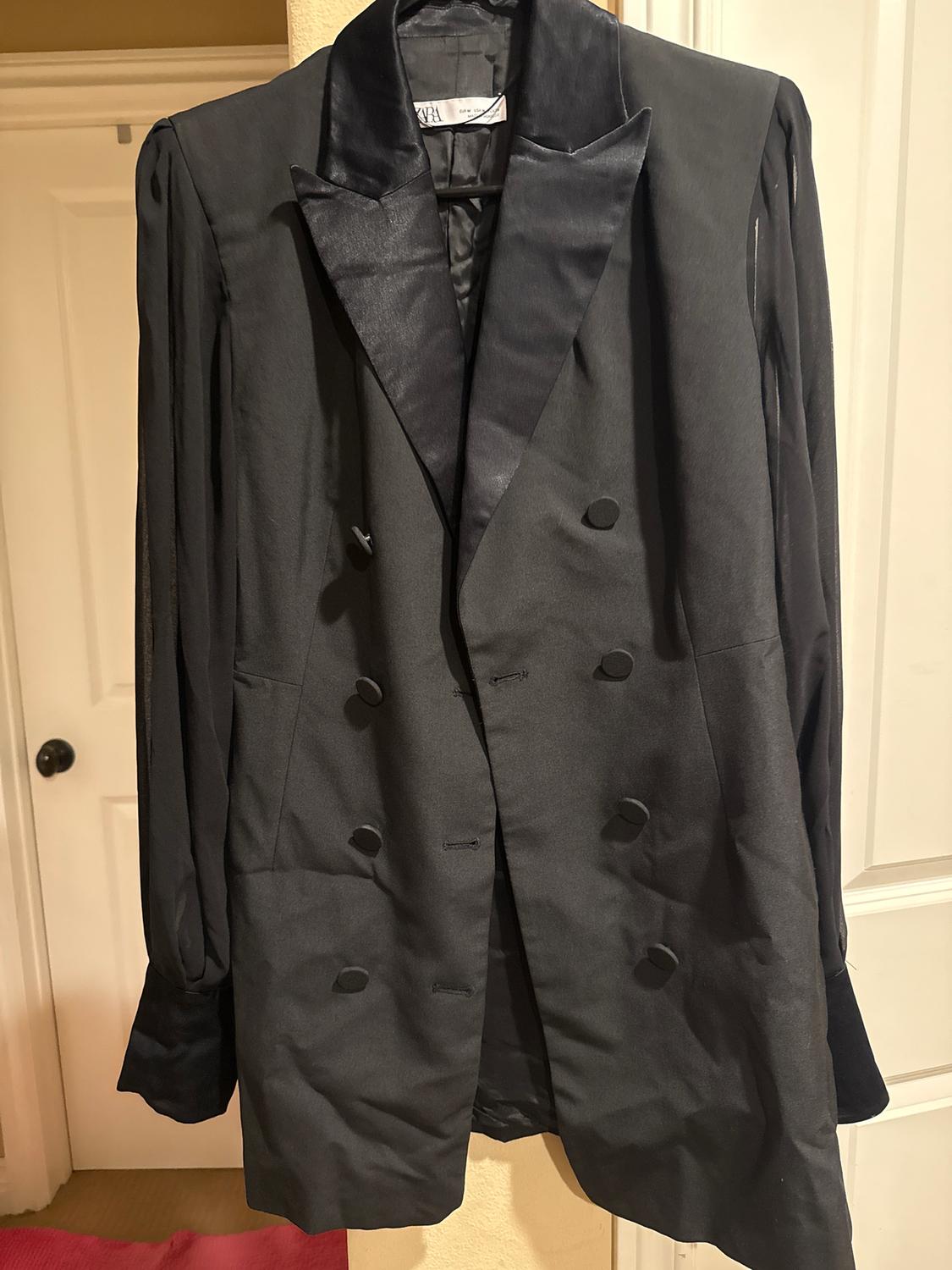 Black double-breasted coat with satin lapels hanging on a hanger, white door background.