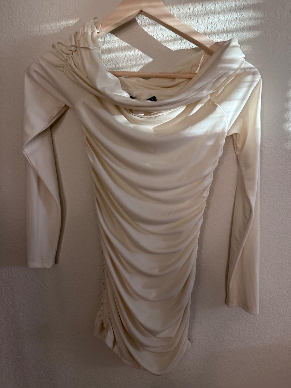 Beige draped long-sleeved dress hanging on a wooden hanger, illuminated by natural light on a light-coloured wall.