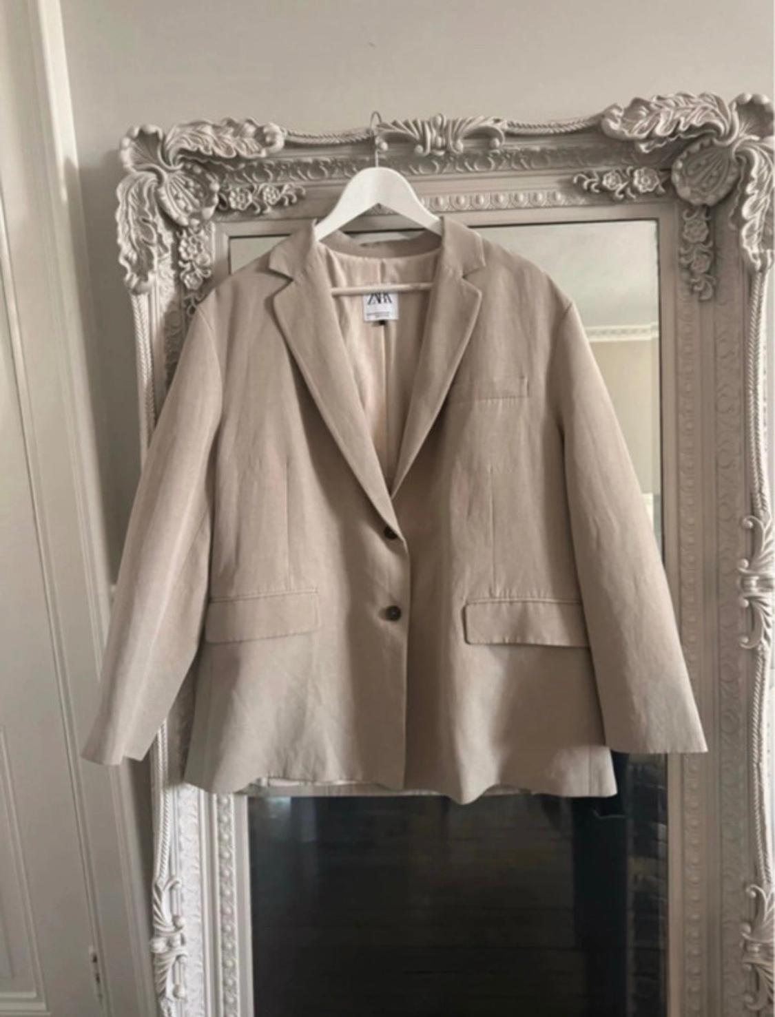 Beige blazer hanging on a white coat hanger in front of a classic mirror with an ornate frame. The inside label reads ZARA.