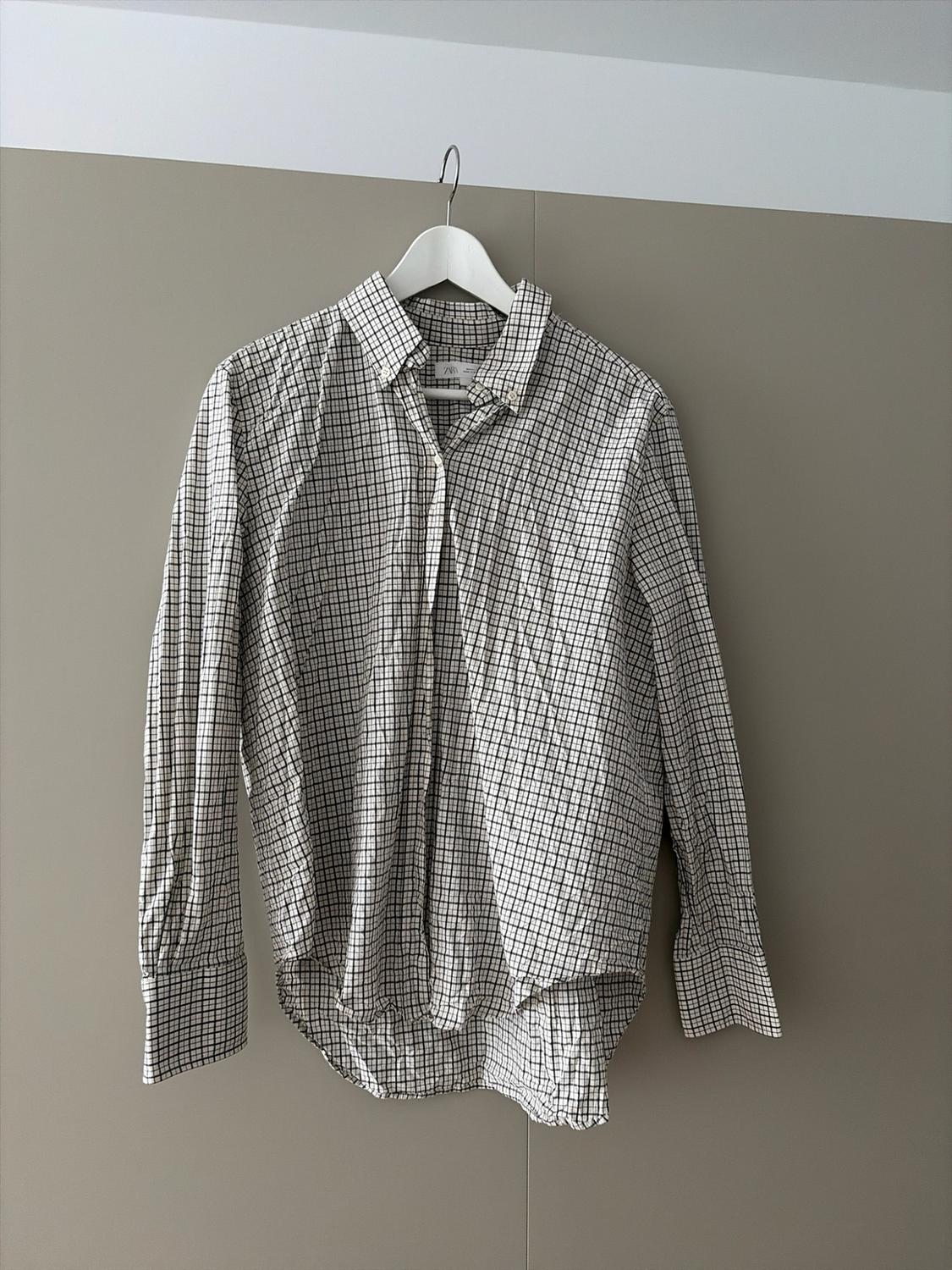 Black and white checked long-sleeved shirt hanging on a white hanger against a beige wall. ZARA label visible.