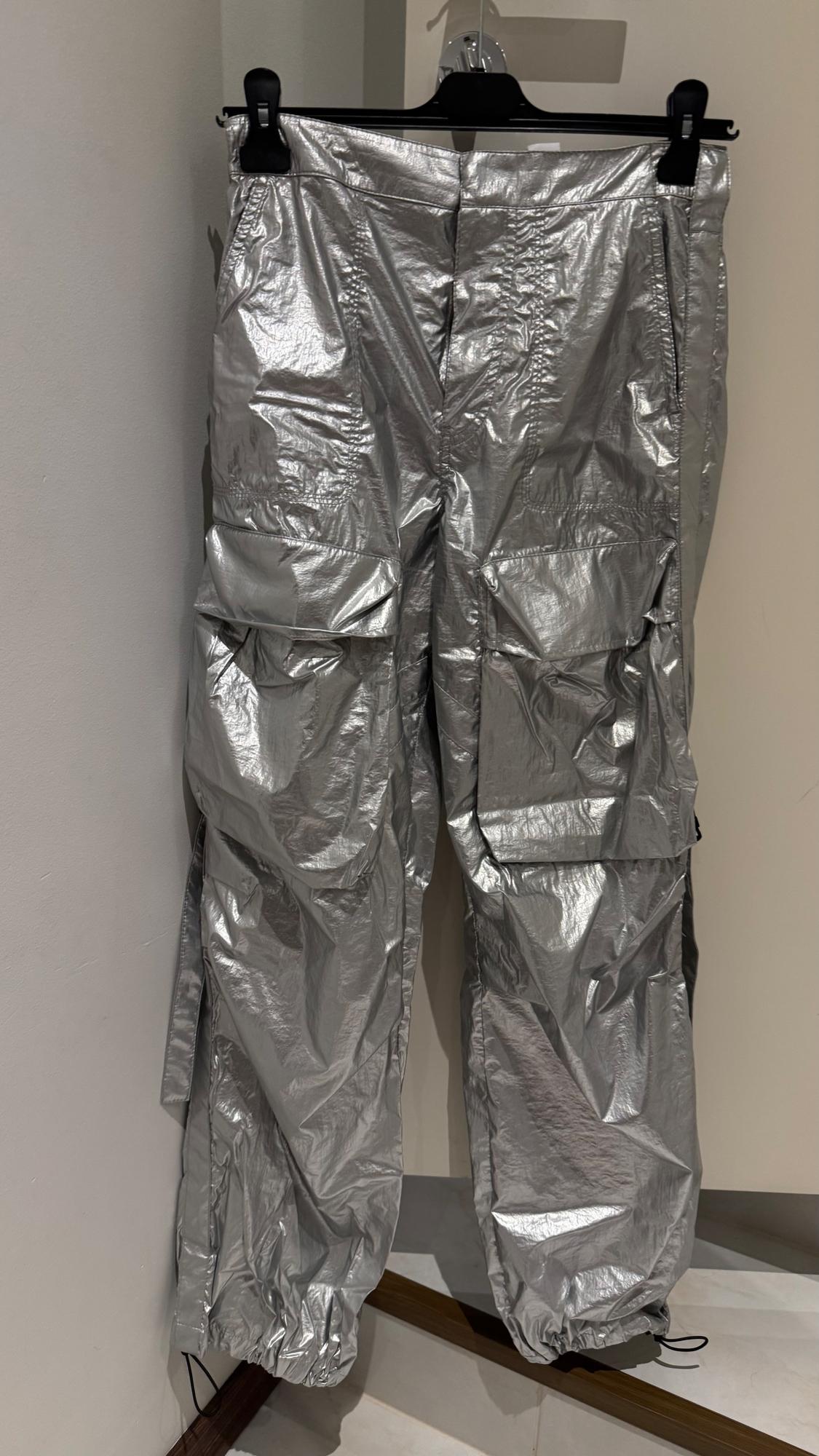 Shiny silver trousers hanging on a black hanger, with large pockets and a wrinkled texture, against a white background.