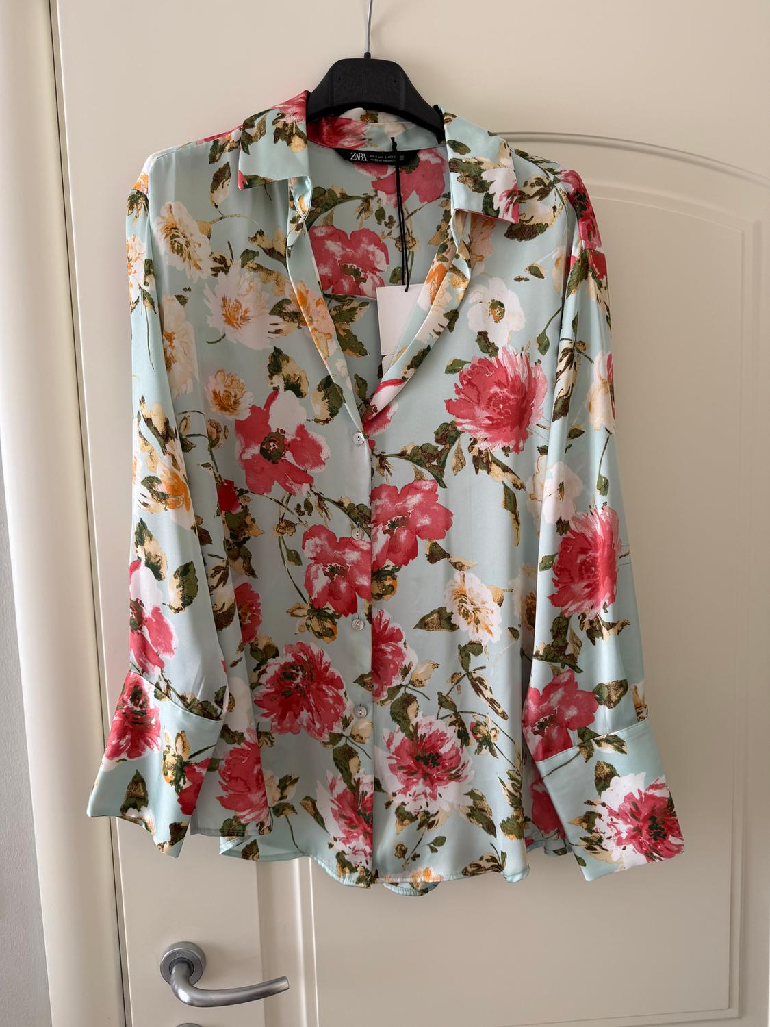Soft fabric blouse with a floral print in pink and green tones, hanging on a black hanger over a white door.