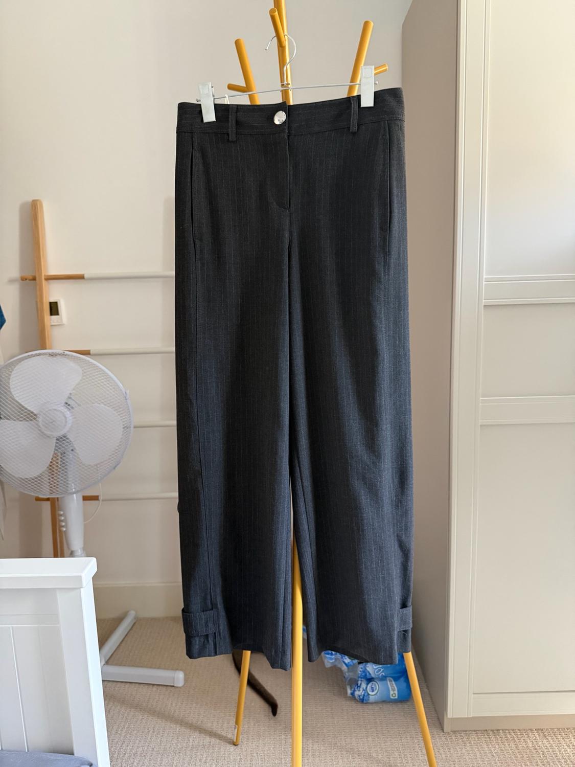 Dark grey pinstriped wide-leg trousers hanging on a yellow coat rack in a room with a fan and a white wardrobe.