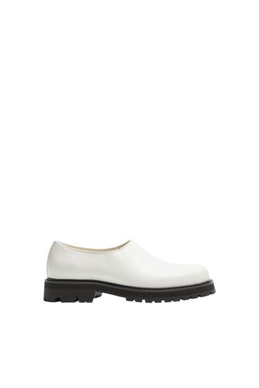 White shoe with a thick black sole on a white background. Minimalist and modern design, with no visible laces or embellishments.