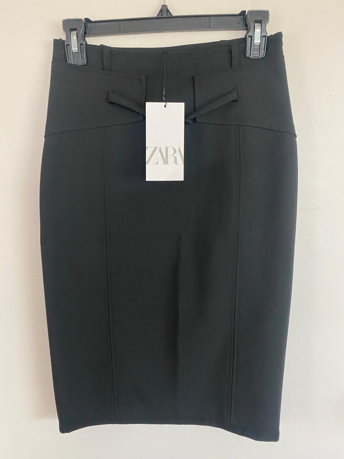 Black skirt hanging on a hanger on a white wall. It has vertical seams and a label with the text ZARA.