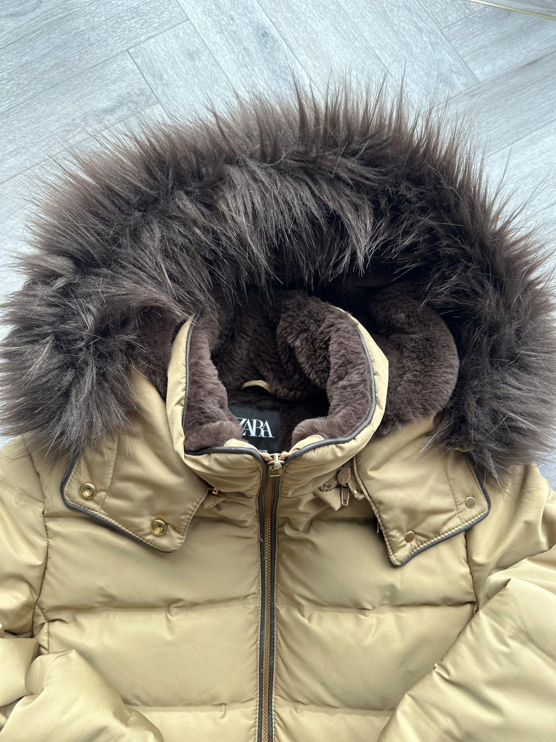 Beige padded jacket with brown faux fur hood, zip fastening and gold buttons on a grey floor. Visible text: ZARA.