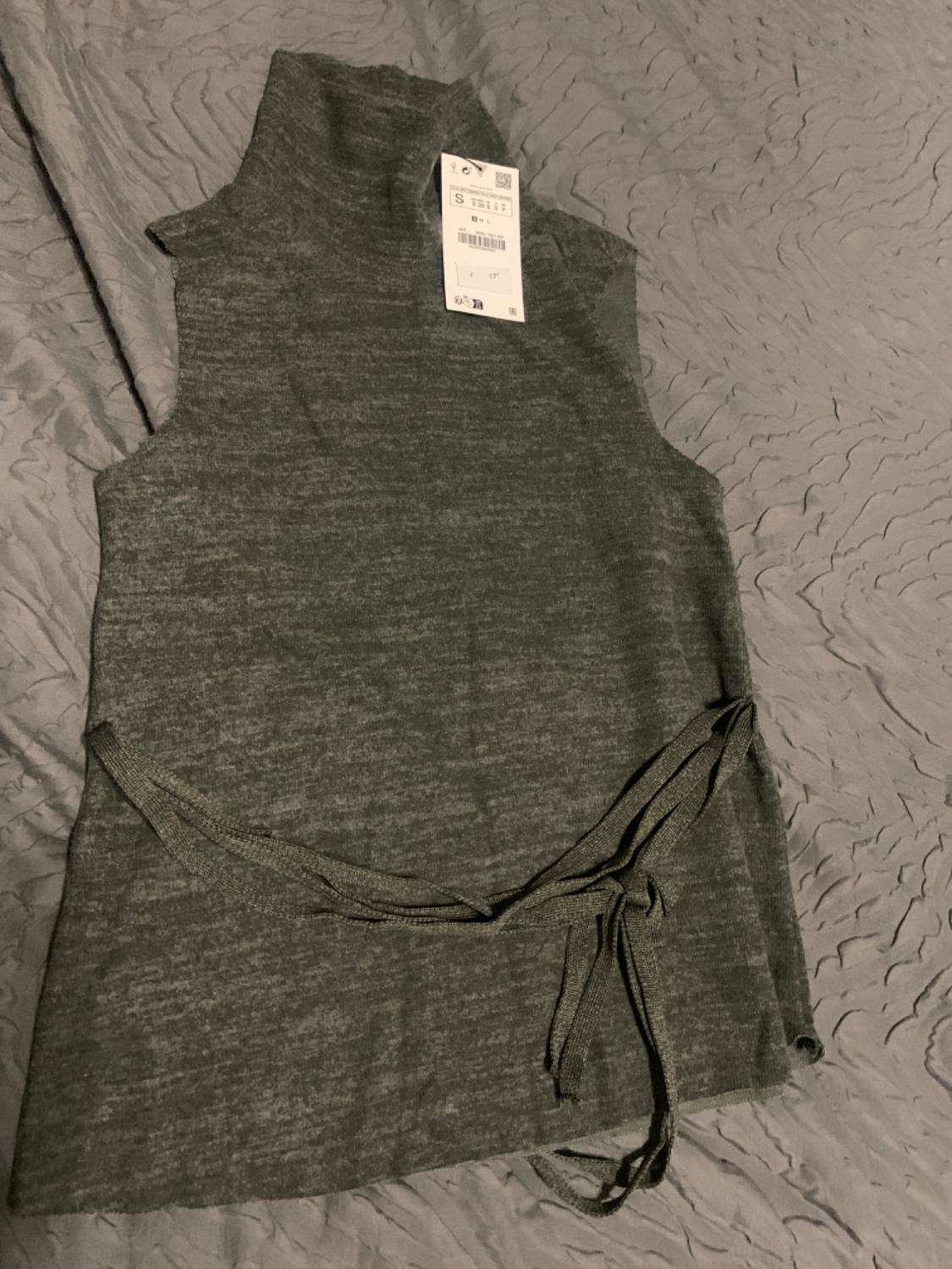 Sleeveless grey gilet with a roll neck and belt, on a wrinkled surface. Visible label with product details.