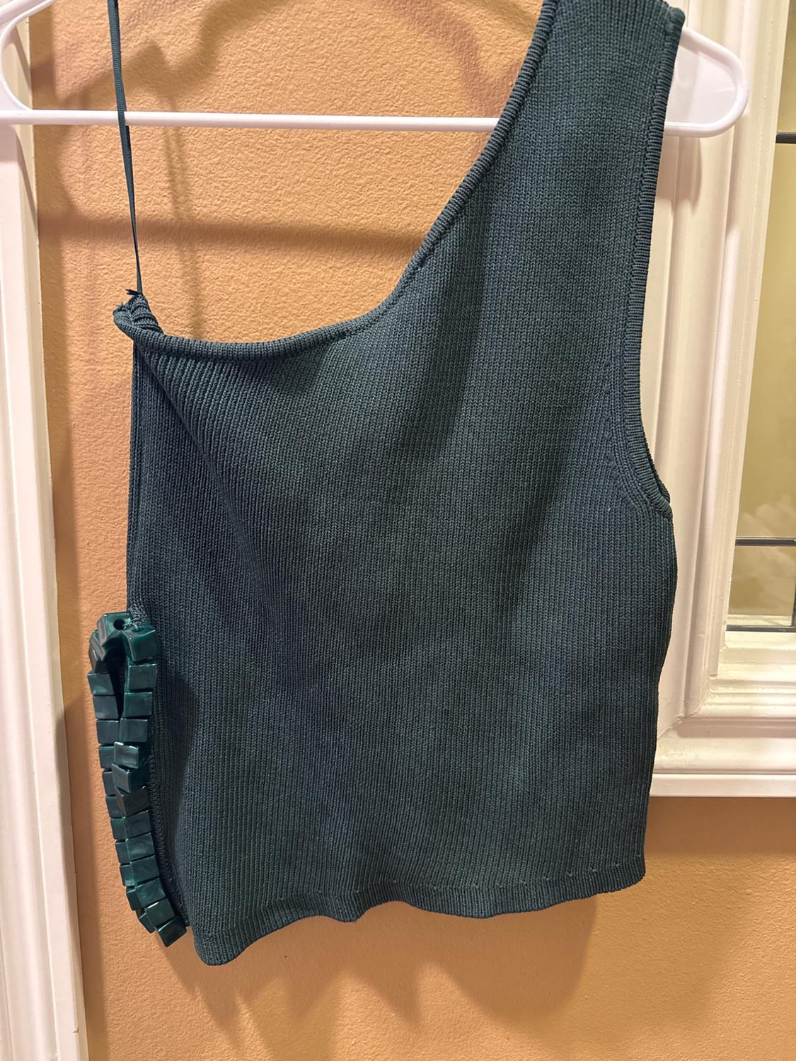 Dark green one-shoulder top hanging on a white hanger, with bead detail on the left side. Beige background.