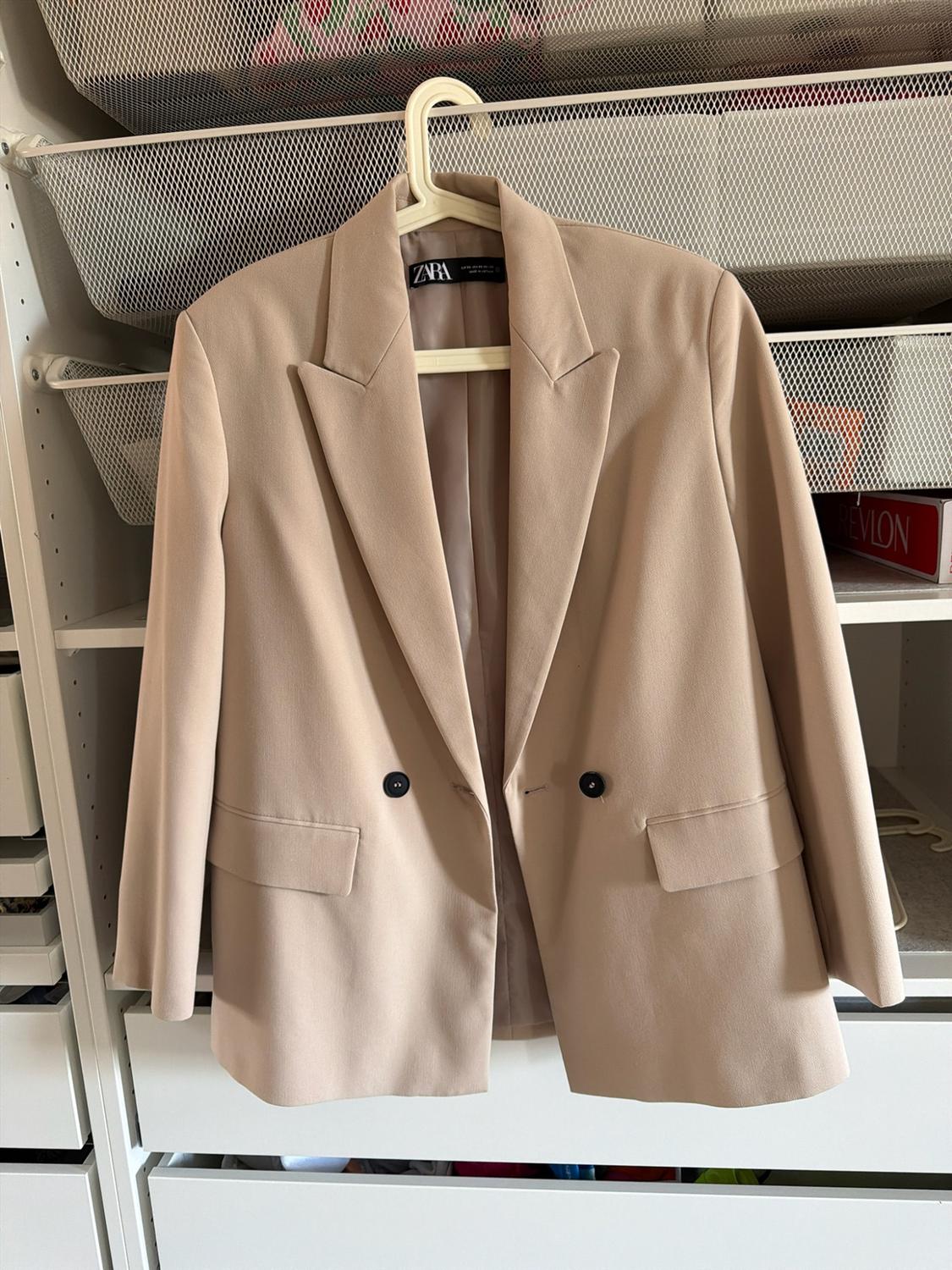 Beige blazer with dark buttons hanging in a white wardrobe. Visible text: ZARA on the label and REVLON in the background.