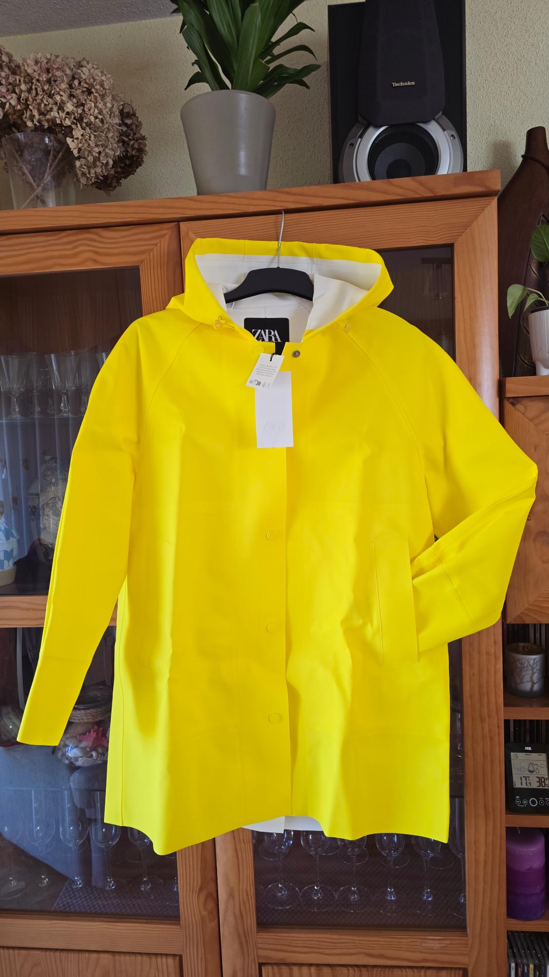 Yellow hooded jacket hanging in a wooden wardrobe. Visible label with the Zara logo. Background with plants and speaker.