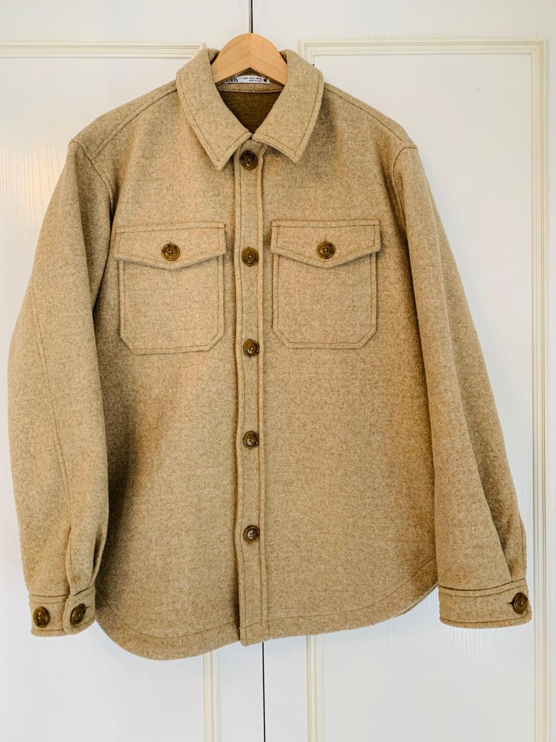 Front view of a beige jacket with front flap pockets and brown buttons.