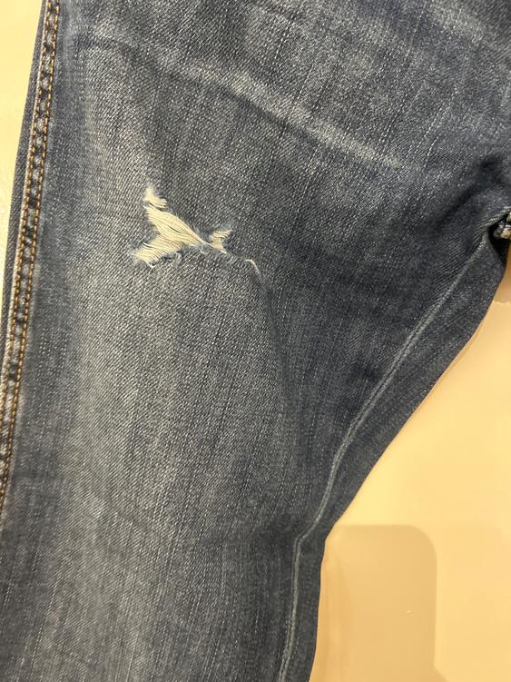 Detail of blue denim trousers with a small tear at the knee, showing visible texture and stitching.