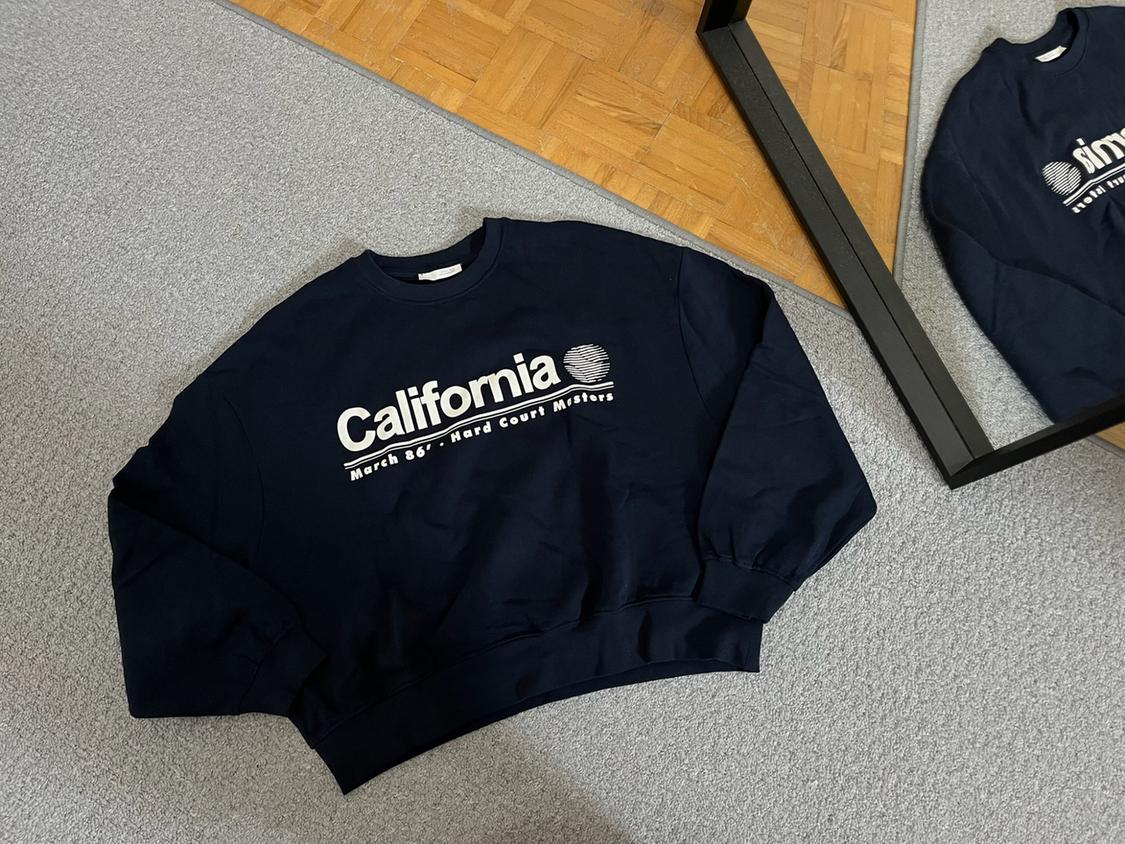 Navy blue sweatshirt on a grey floor with the text 'California March 86' - Hard Court Masters'. A mirror reflects the garment and a wooden floor.