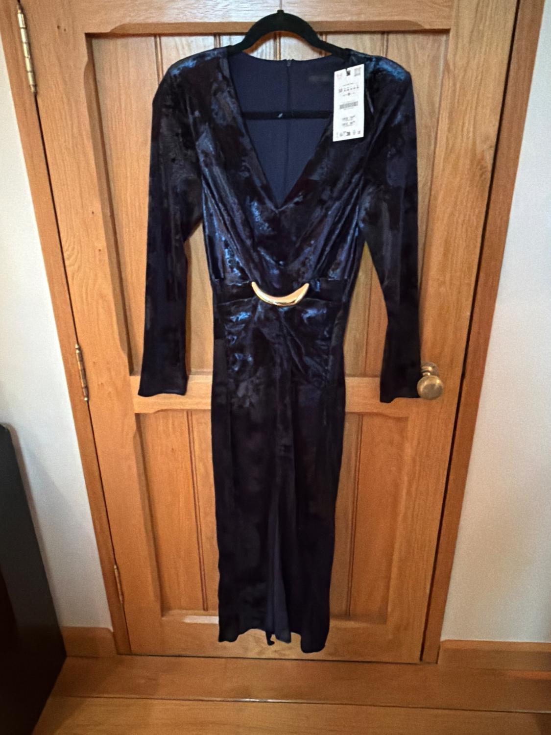 Dark blue velvet maxi dress with a V-neckline and gold belt, hanging on a wooden door. Label visible.