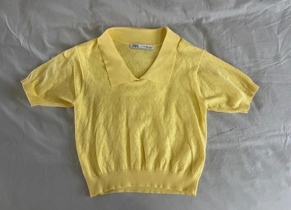 Yellow openwork knitted polo shirt with short sleeves, laid out on a crumpled grey fabric. The inner label reads ZARA.