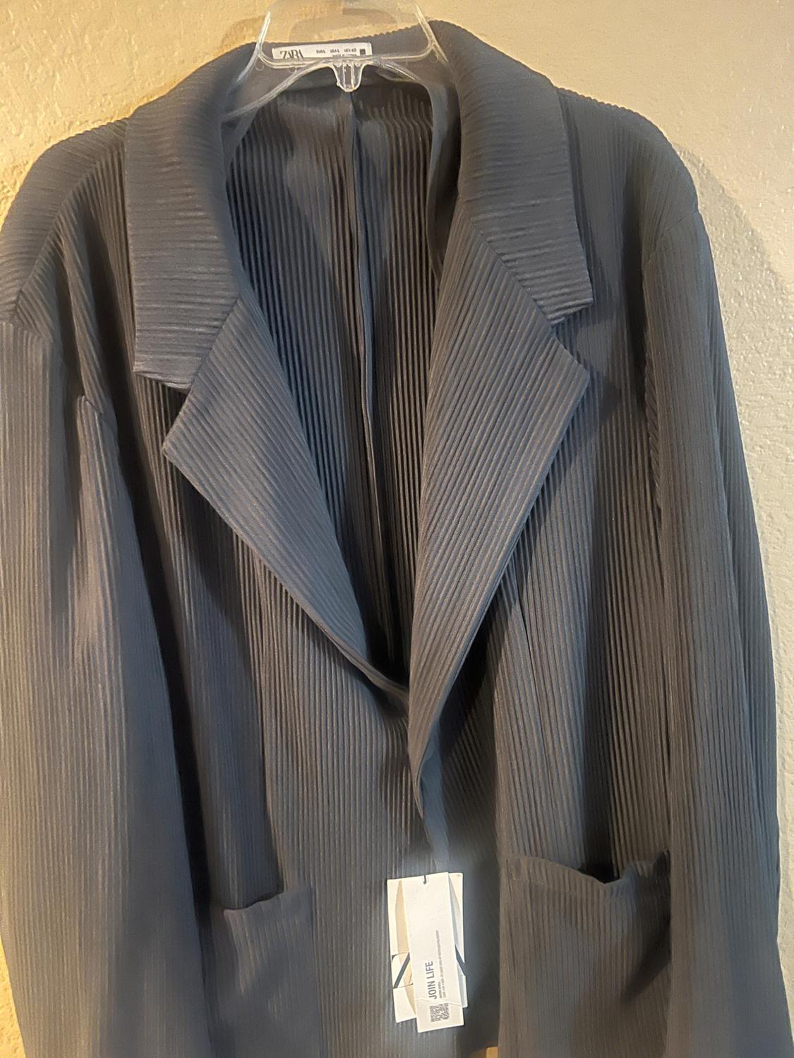 Grey pleated fabric jacket hanging on a hanger, with a visible label including the text "Join Life" and a QR code.