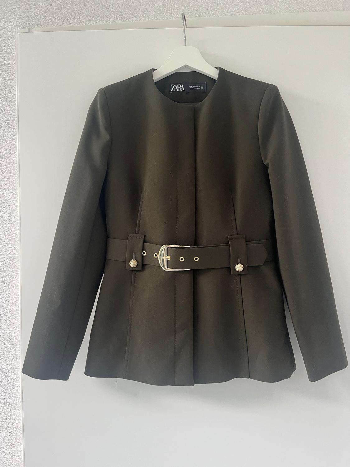 Olive green jacket with belt and gold buckle, hanging on a white hanger. Visible label: ZARA. White background.