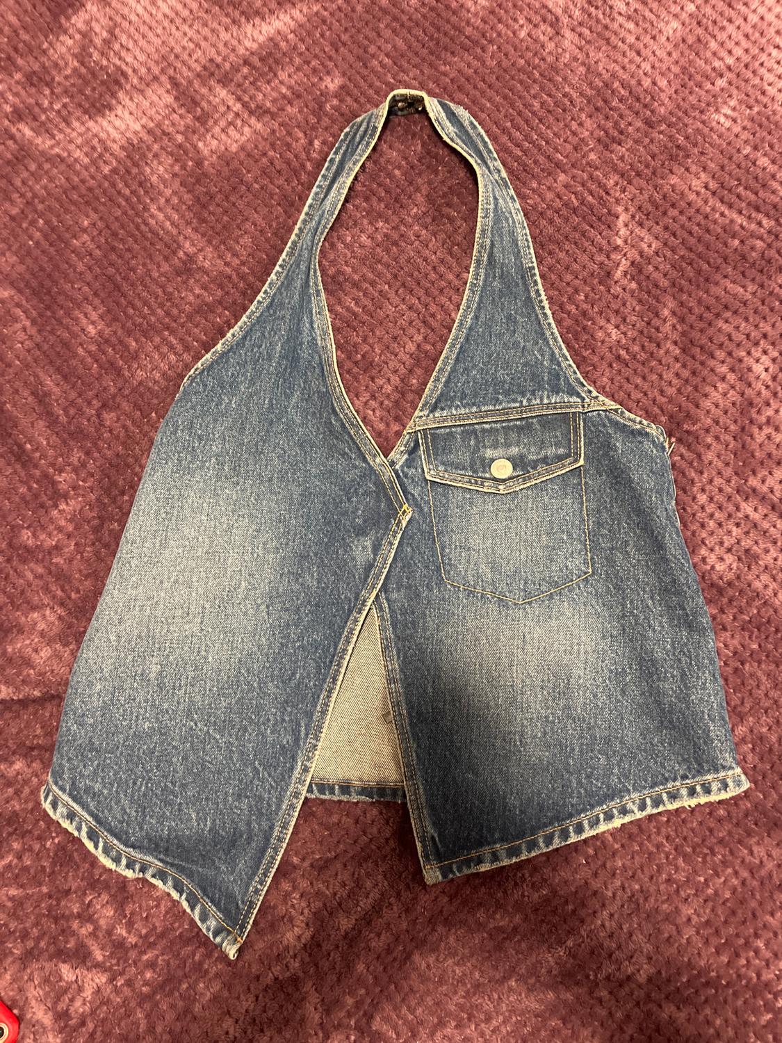 Blue denim waistcoat with front pocket and button fastening, on a textured burgundy surface.