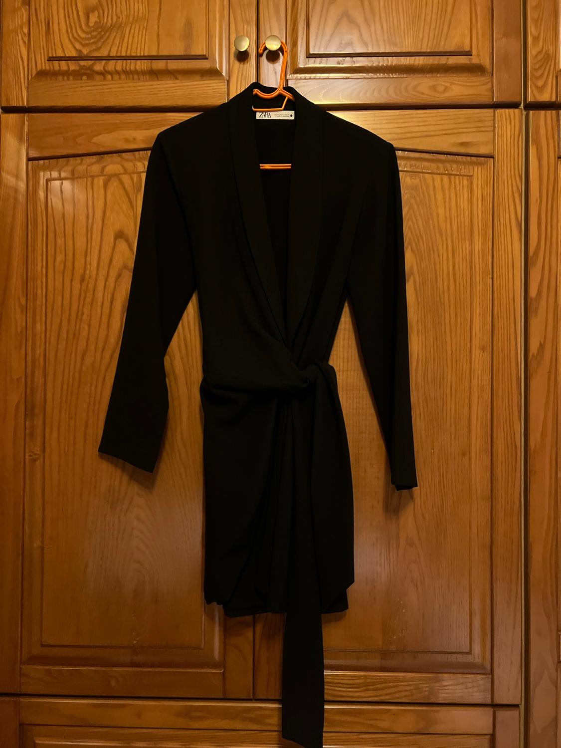 Black long-sleeve wrap dress with a knot detail, hanging on an orange hanger in a wooden wardrobe. ZARA label.