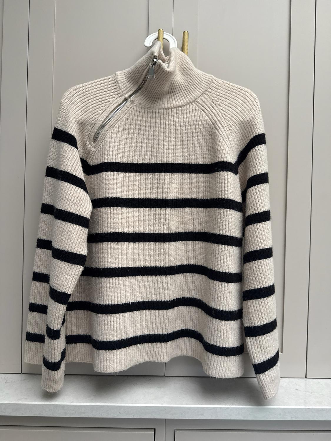 Beige knitted jumper with black stripes and a high neck with a zip, hanging in a light grey wardrobe.