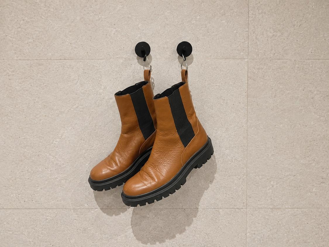 Pair of brown boots with black elasticated sides and thick soles, hanging from two hooks on a light-tiled wall.