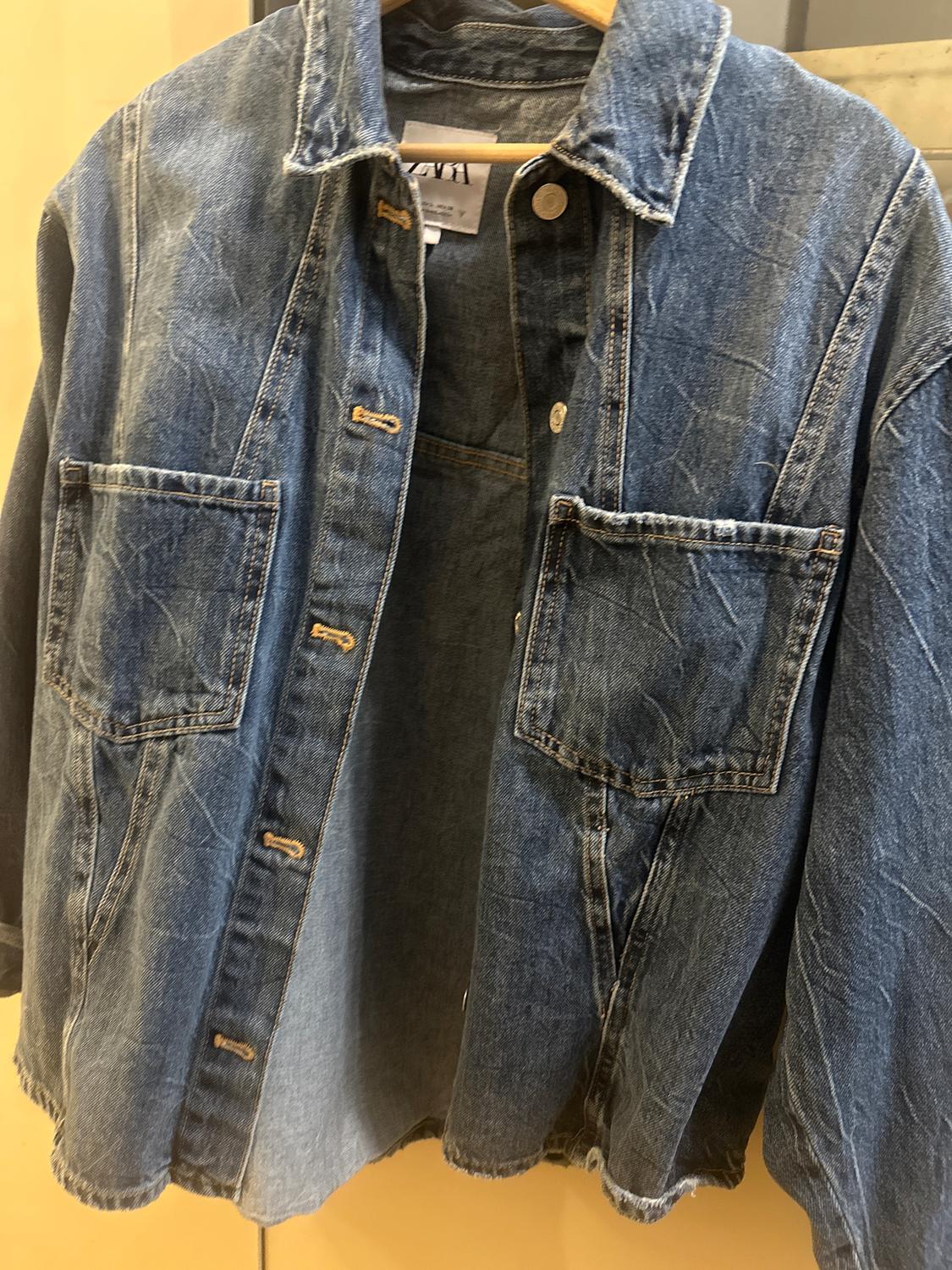 Blue denim jacket hanging on a hanger, with large pockets and metal buttons. Visible label on the collar.