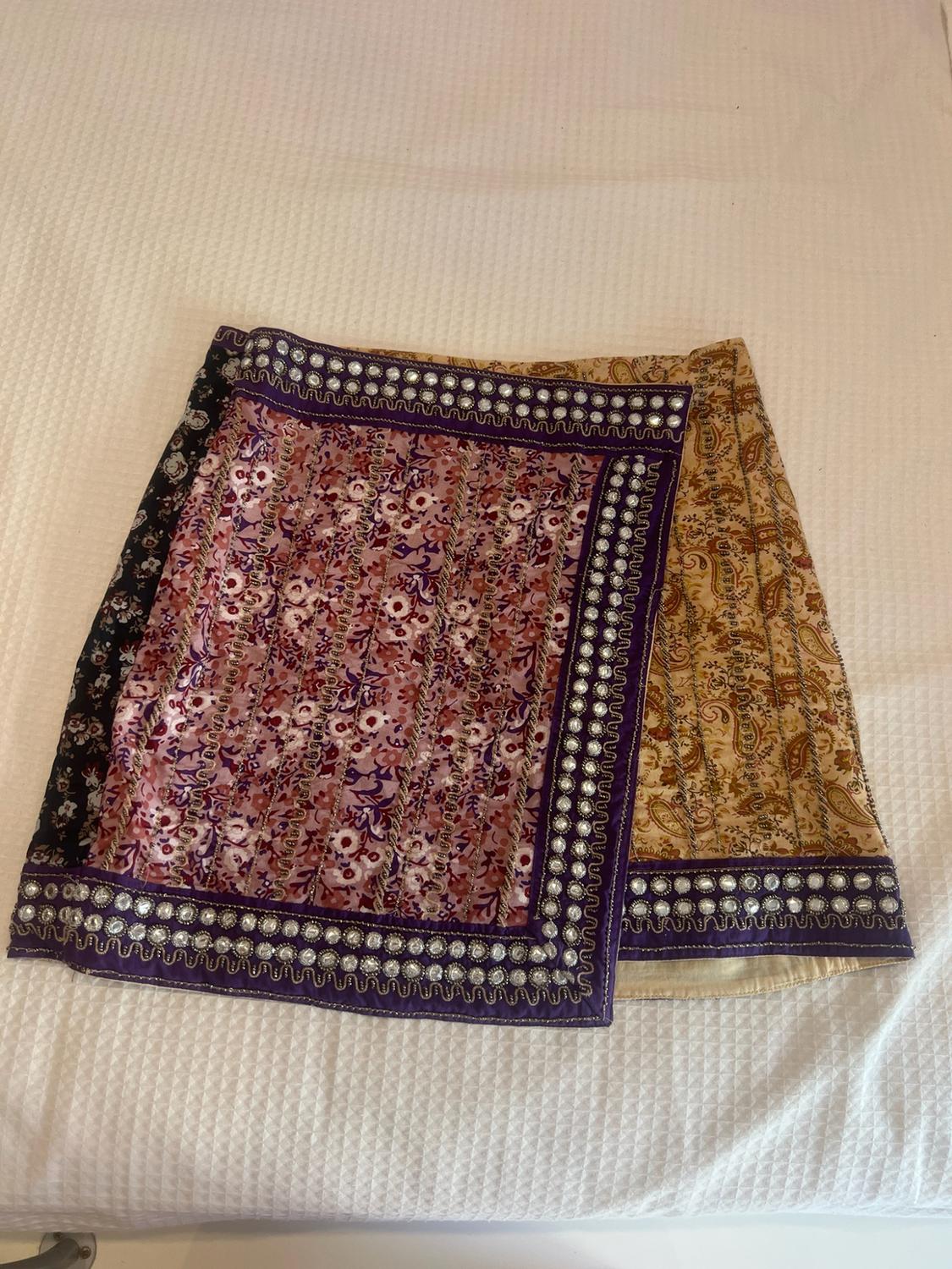 Short wrap-around skirt with a purple and beige floral print, featuring sparkly appliqué edging, set against a textured white background.