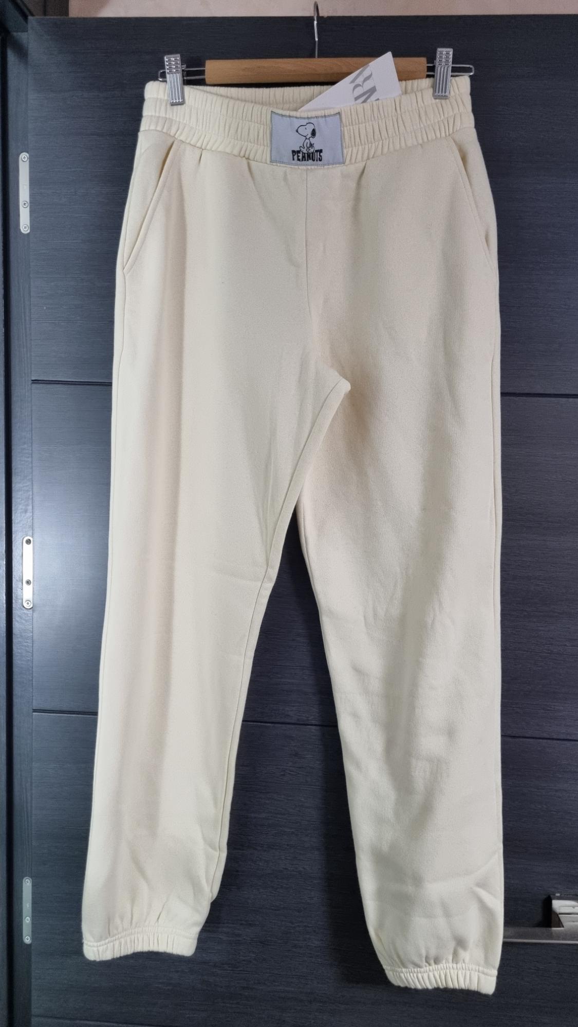Beige trousers hanging on a hanger, with an elasticated waist and a "Peanuts" label featuring Snoopy at the top.
