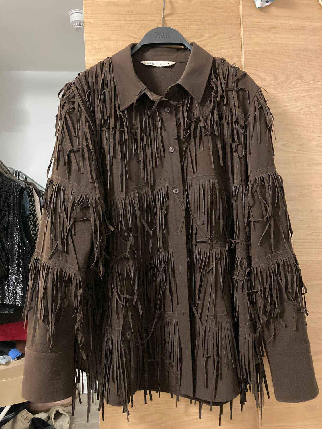 Brown fringe shirt hanging on a black Zara hanger, light wooden background and clothes on the left side.