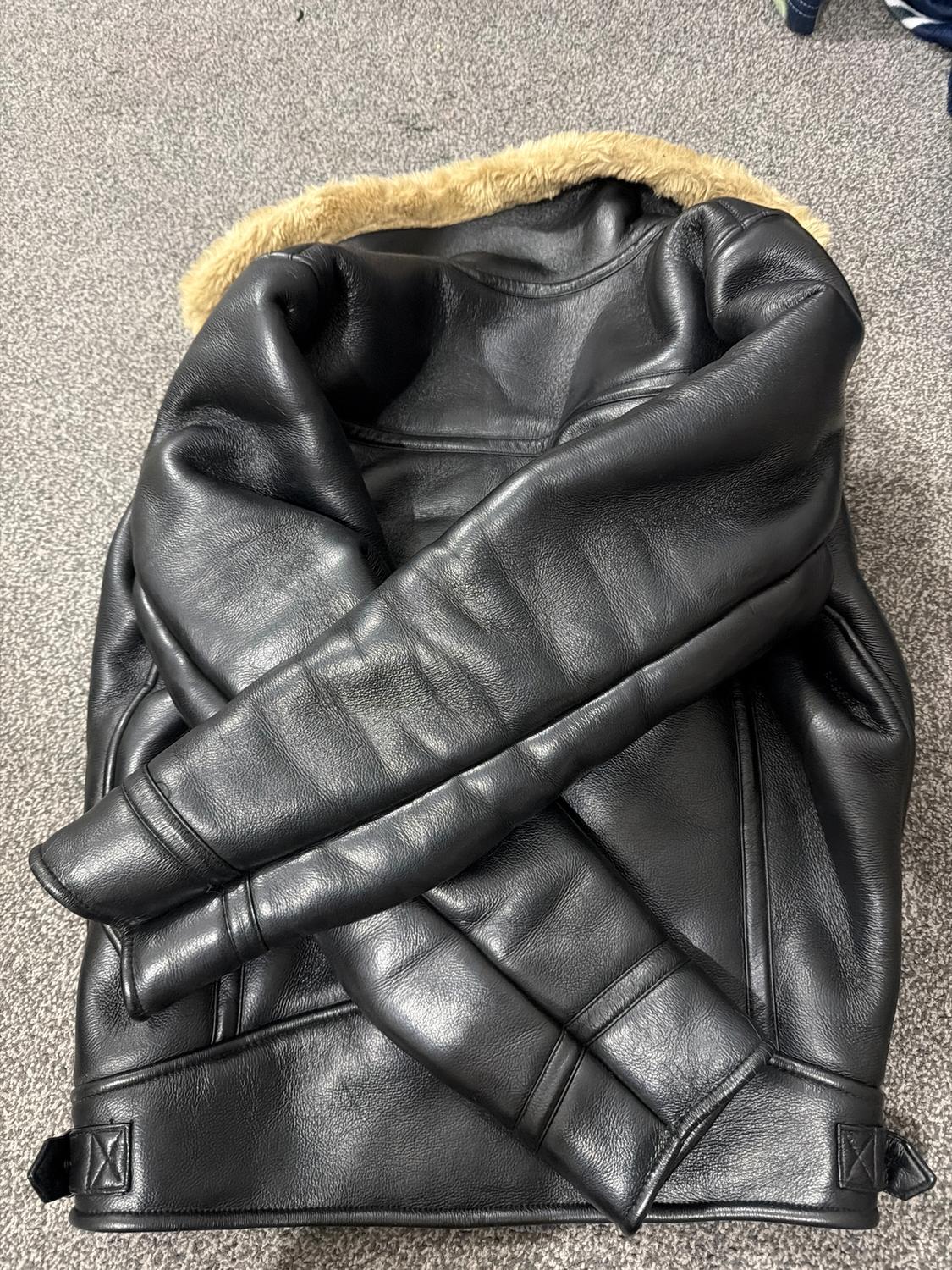 Black leather jacket with a light brown fur lining, placed on a grey rug. Sleeves crossed at the front.
